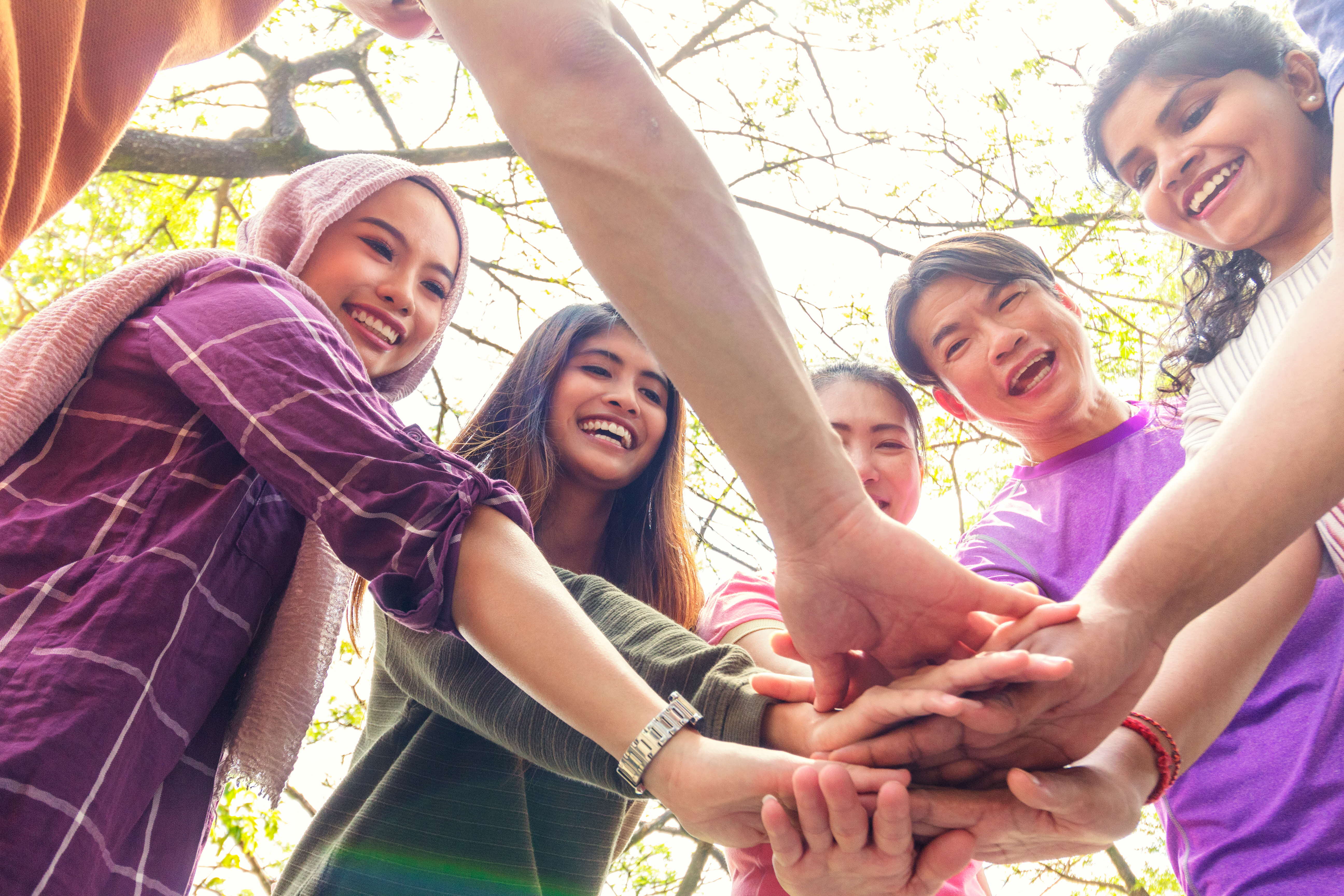 Group of smiling youths in a circle stacking hands together outdoors under a sunny sky.